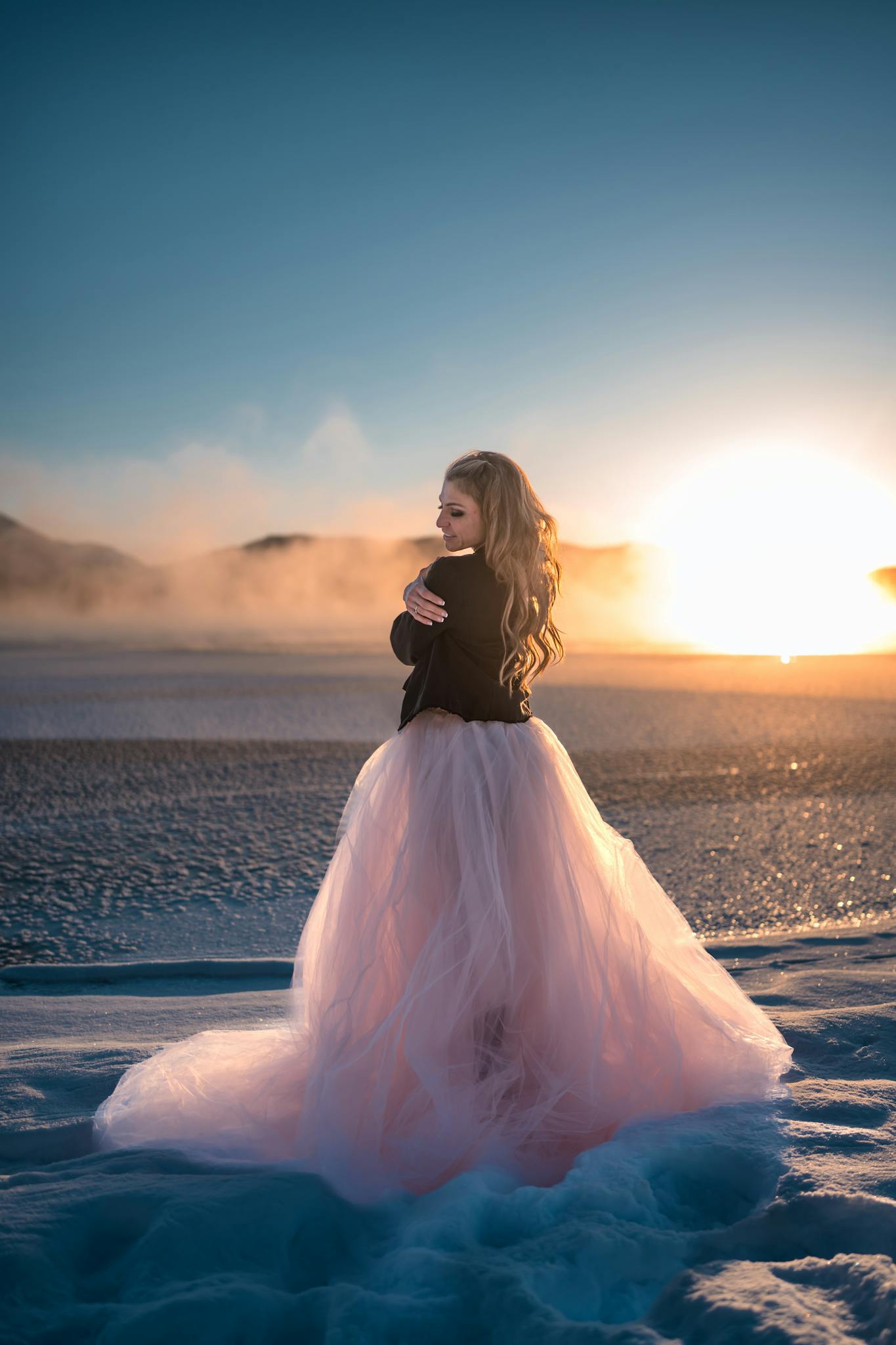 An elegant woman in a pink tulle dress stands on a beach at sunset, exuding beauty and grace.